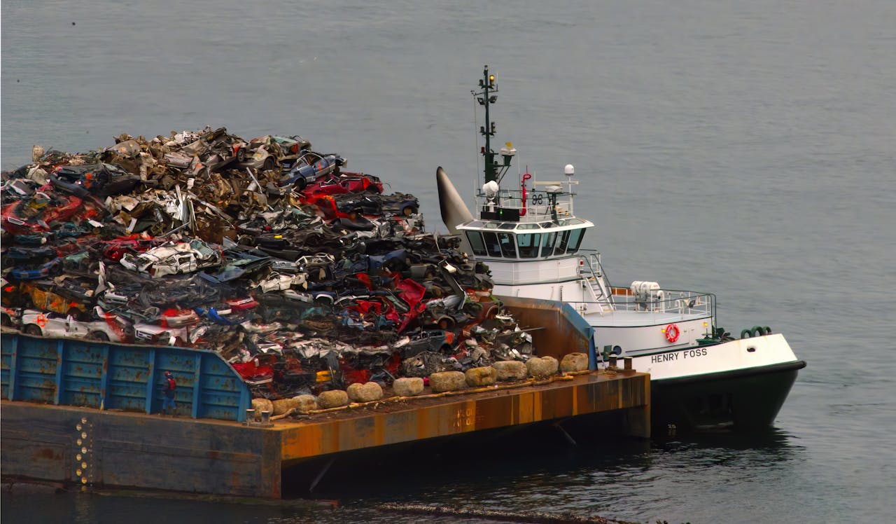 A barge loaded with crushed cars and scrap metal transports cargo over the ocean with a tugboat.